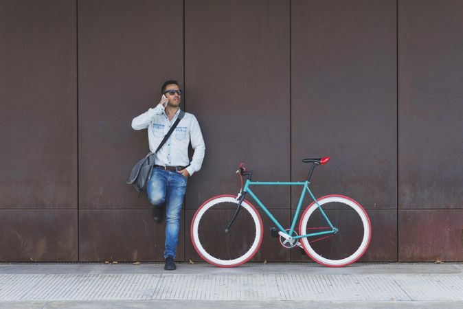 Male in sunglasses standing with red and green bicycle leaning on wall and using phone