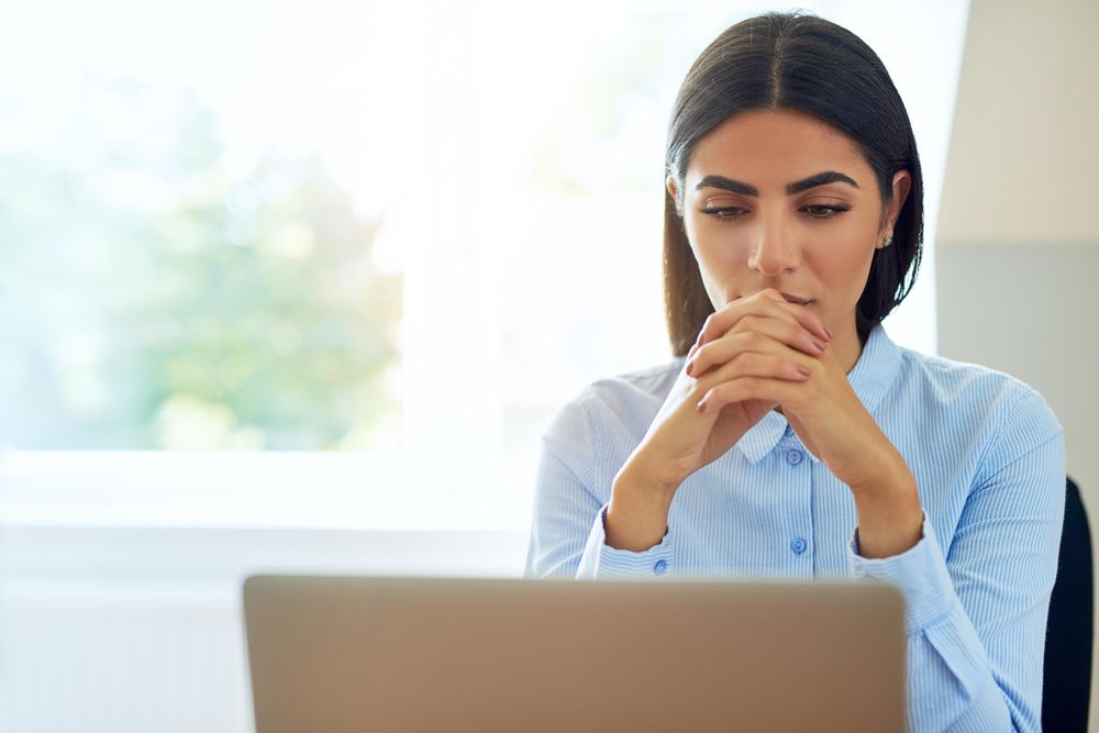 Pensive female sitting at her laptop reading screen with hands clasped