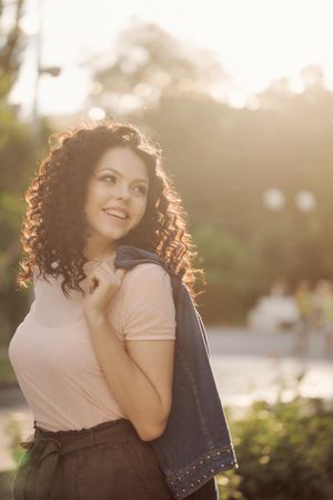 Smiling young woman looks to her side outside while holding coat over shoulder