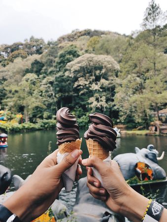 Couple Sharing Chocolate Ice Cream Cones by a Serene Lake with Lush Greenery
