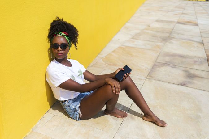 Portrait of Black woman in t-shirt sitting against yellow wall with phone