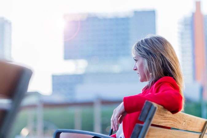 Elegant woman in red jacket sitting on a bench outdoors while looking away in sunny day