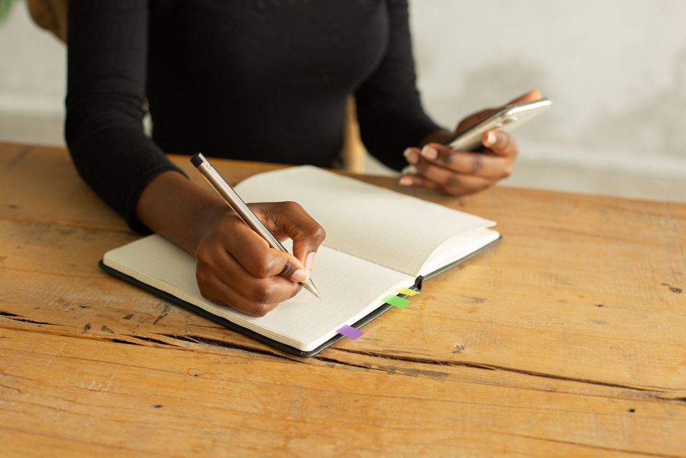 Close-up shot of woman writing in a notebook while looking at smartphone