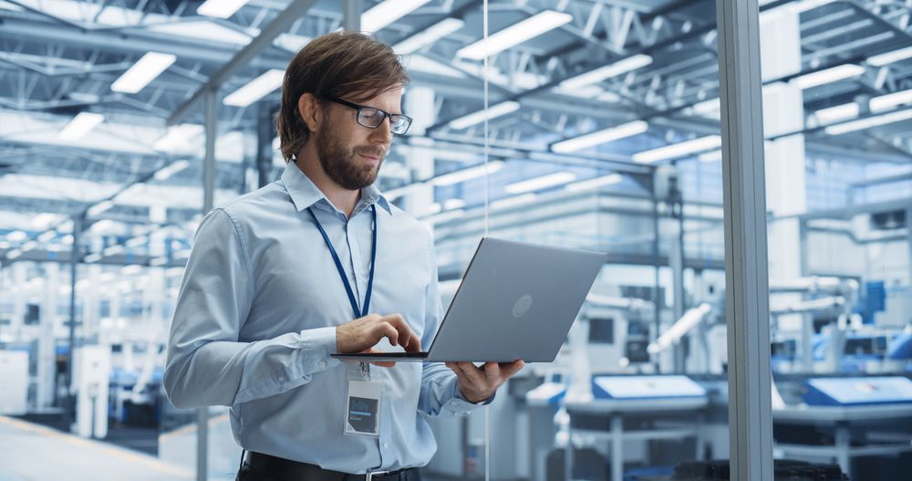 Portrait of a Handsome Engineer with Glasses Using Laptop Computer and Looking Around a Factory Facility with Equipment Producing Modern Electronic Components for Different Industries