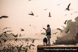 Side view of an Indian man in kurta standing by riverside with a dog while birds flying 5kMX3b