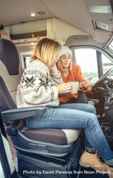 Female friends sitting in parked van having coffee and checking social media on phone, vertical 4BNwB4