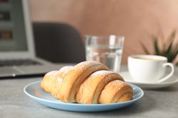 Blue plate with croissant, coffee and water next to computer on grey table, side view, close up