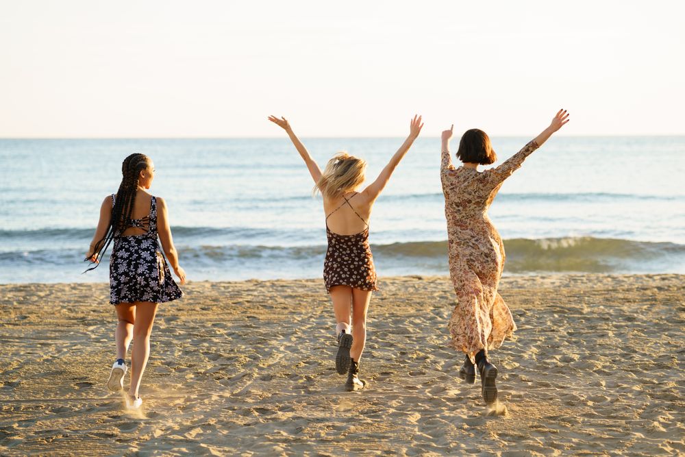 Backs of three happy women skipping towards the coast on beach with arms up