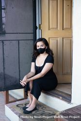 Woman sitting on front door steps of her house wearing mask and looking at camera bE9oAb