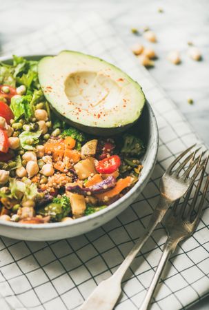Close up of lunch bowl with avocado, sprouted grains, vegetables, with forks on checkered napkin