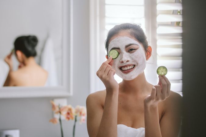 Smiling young woman with face pack holding slice of cucumber to eyes