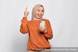 Muslim woman smiling holding smart phone and pointing fingers up with a bright idea 5RG7J4