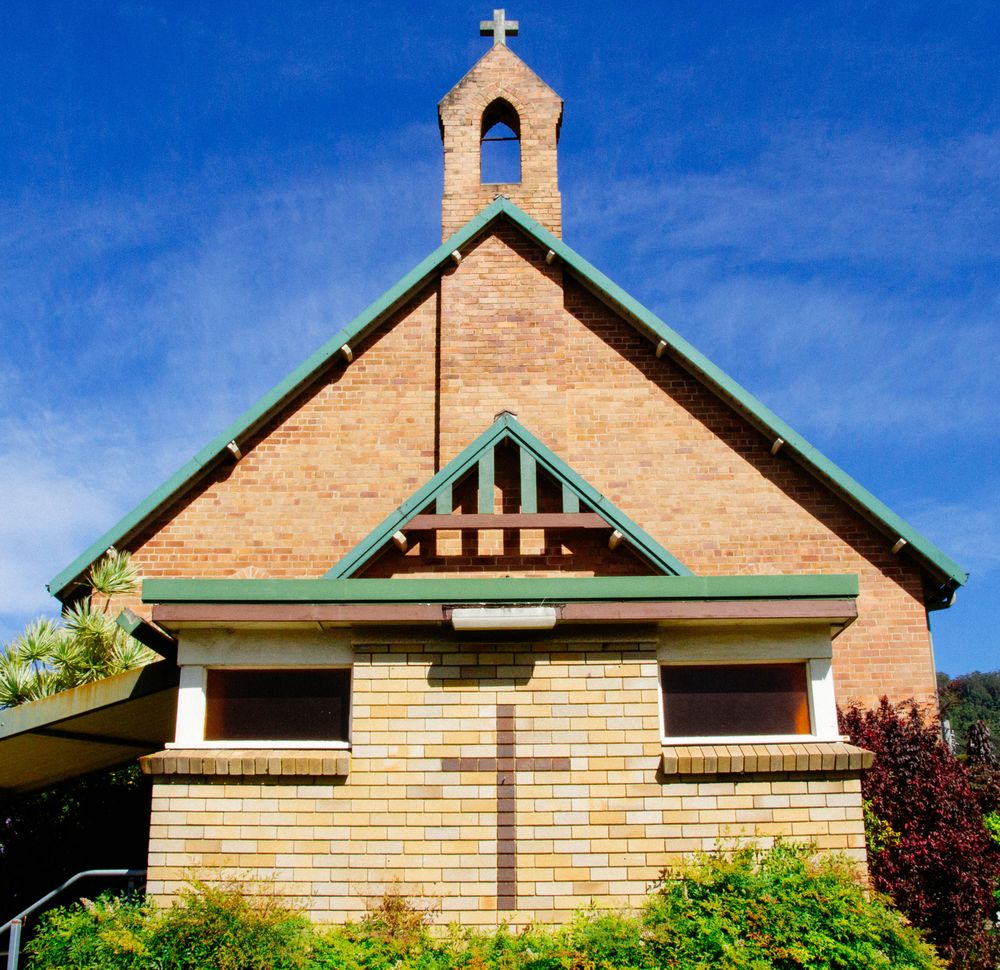 Front View Of A Brick Church With Green Roof And Cross Against A Clear Blue Sky