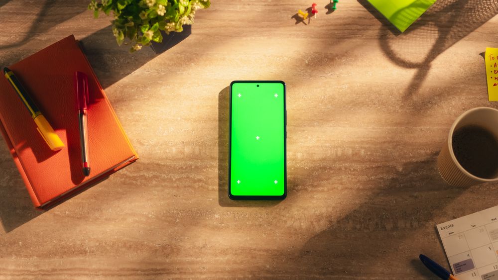 Top Down View of a Smartphone with Mock Up Green Screen Display. Cellphone Device Lying on a Wooden Table in a Creative Office. Template for Online Digital Marketing, Social Media.