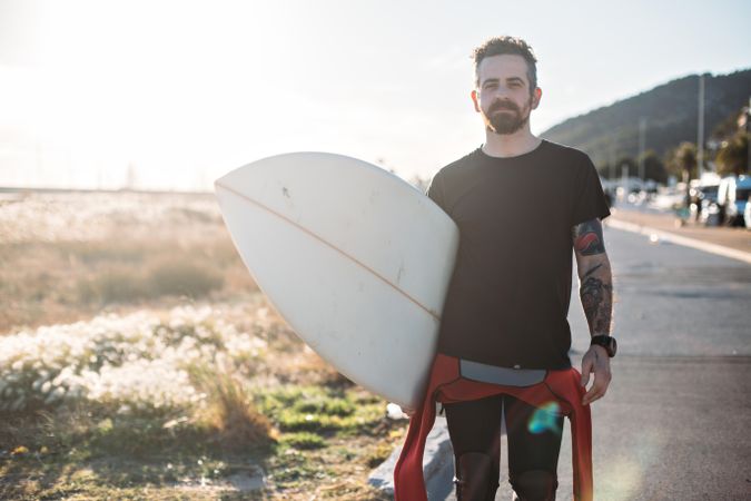 Tattooed man standing on road holding surfboard