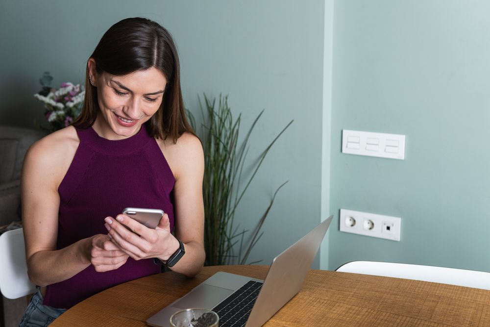 Smiling woman using smartphone and sitting at table with laptop