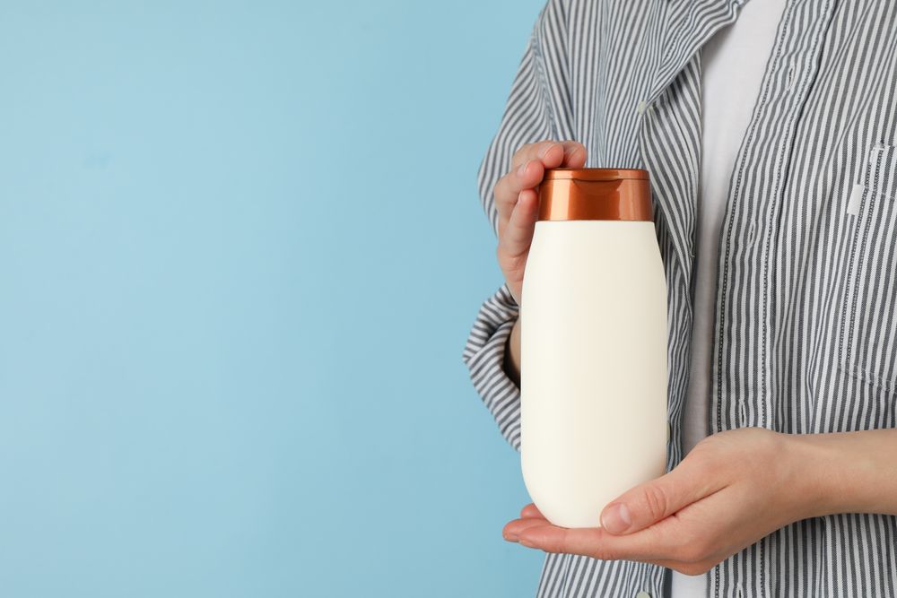 Woman holds blank shampoo bottle on blue background, space for text