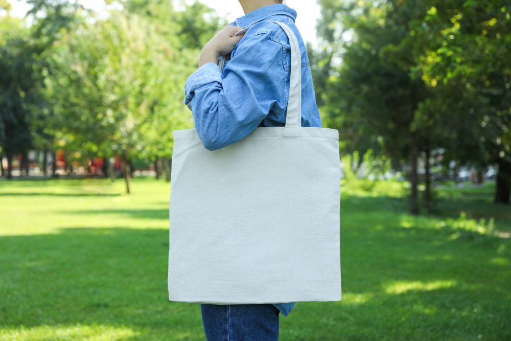 Young woman holding tote bag against greenery background, empty space