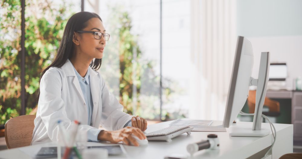 Young Female Doctor Wearing Coat, Working on Personal Computer in Her Office.