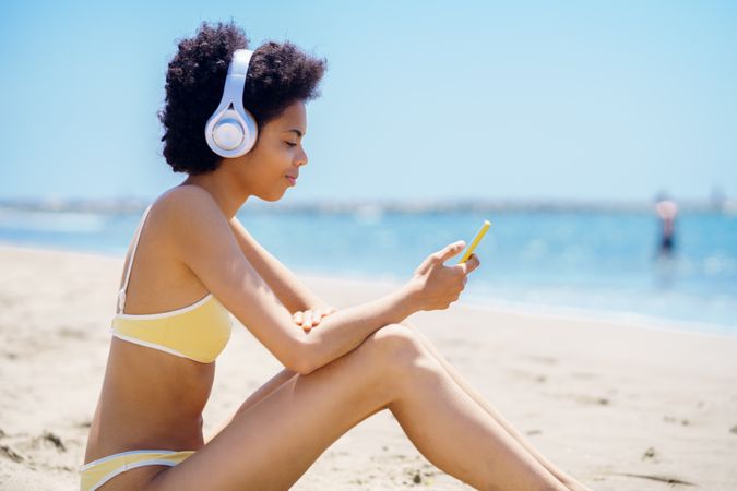Woman in yellow bikini sitting on beach and looking at phone
