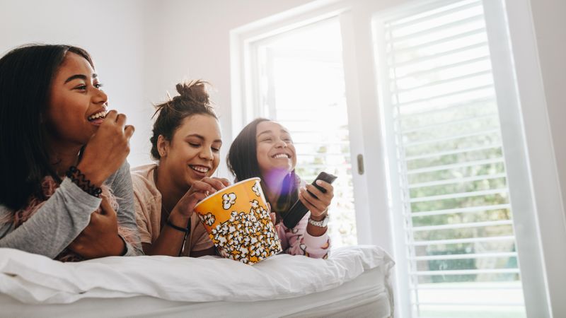 Group of young women lying on bed eating popcorn from a tub watching TV