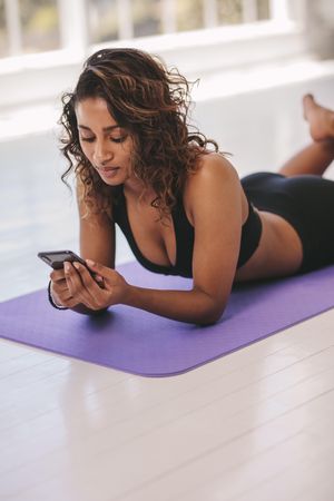 Fit woman lying on fitness mat and watching exercise videos on smart phone
