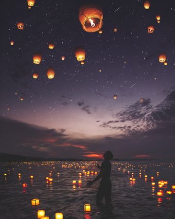 Silhouette of man standing on body of water during night time with lit lanterns in sky