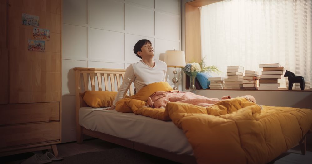 Korean Young Man Wakes Up in his Bed, Sun Shines on Him, He Greets a New Sunny Day at Home and Stretches.