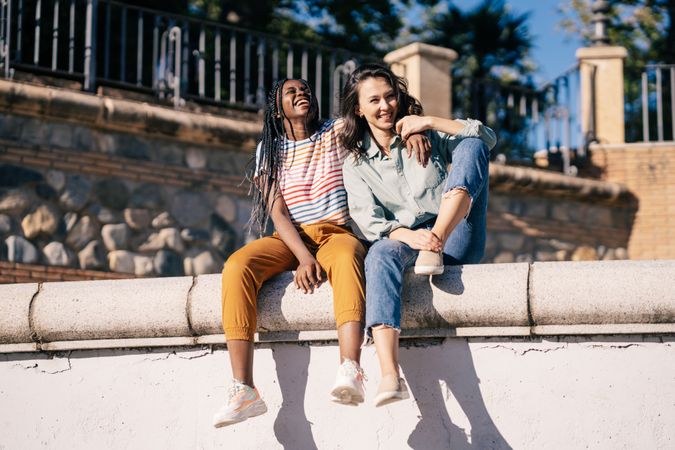 Close female friends sitting on outdoor wall on sunny day
