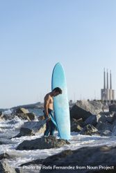 Male surfer with blue board standing in the sea around a rocky beach 569XN0