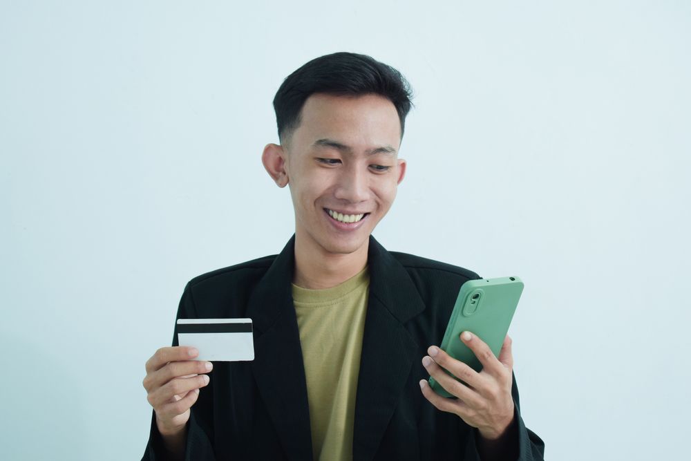 Joyful Asian businessman in suit holding blank credit card and smartphone