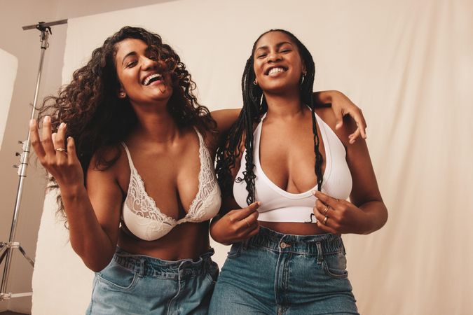 Two happy female friends wearing denim jeans and bras against studio background
