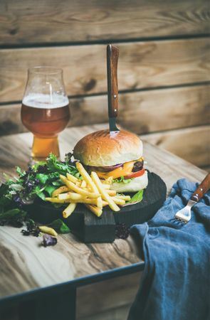 Classic hamburger skewered with knife with fries and beer at wooden restaurant table