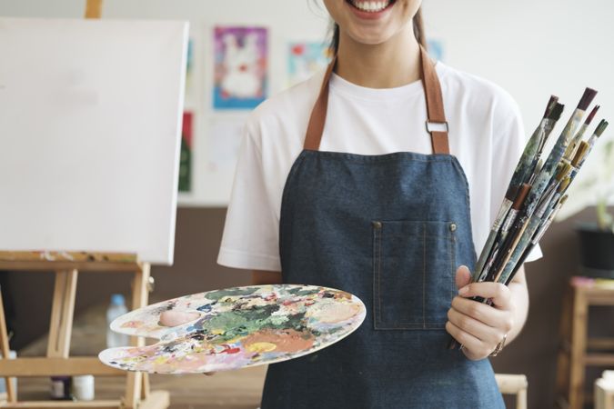 A close-up of the artist hands wearing an apron smeared with paint. Clutching many brushes and paintbrushes.