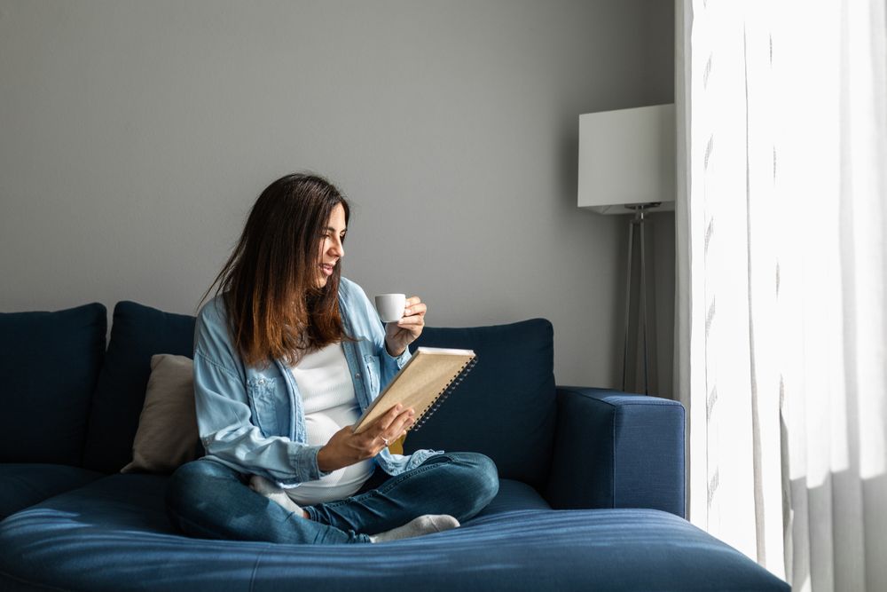 Cheerful pregnant woman drinking beverage on sofa at home