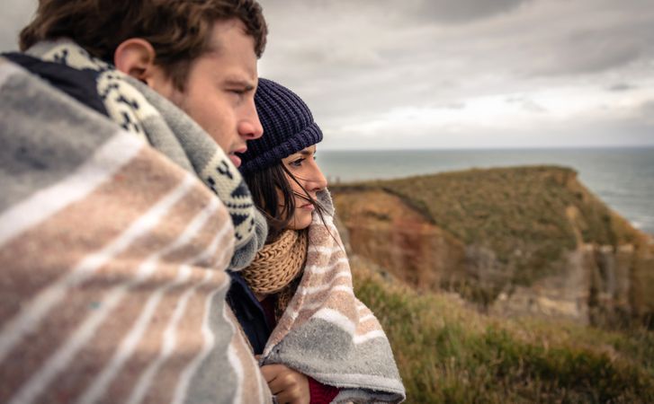 Side view of loving couple wrapped in blanket on a windy day on a cliff above the coast