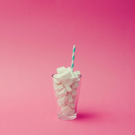 Glass with blue straw filled with sugar cubes on pink background