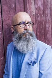 Front view of a mature trendy bearded hipster leaning on a vintage red door outdoors while looking away