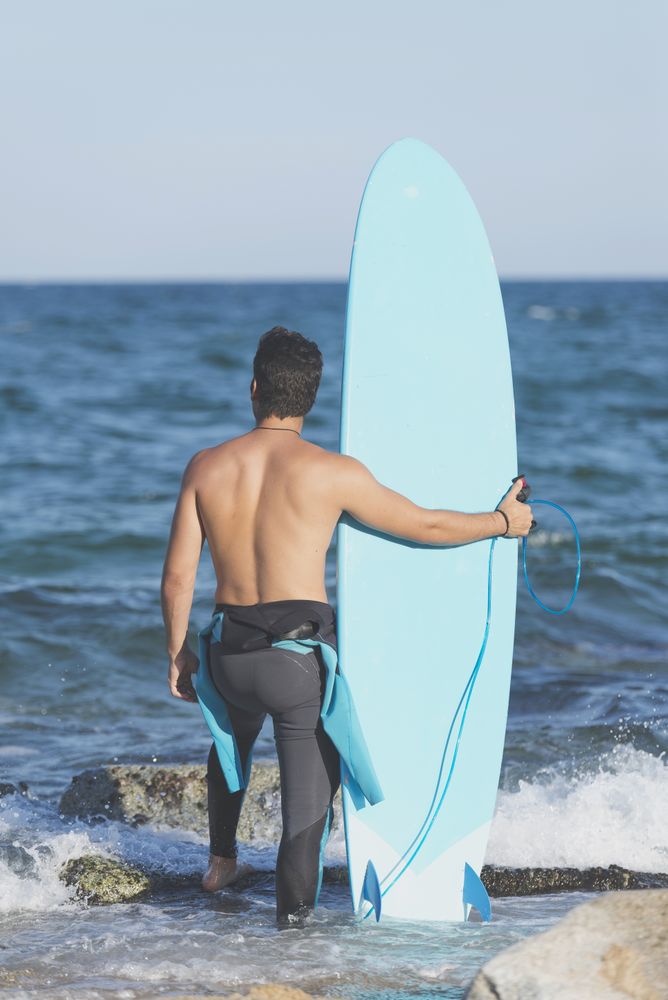 Male surfer with blue board enjoying the view of the water from rocky shore