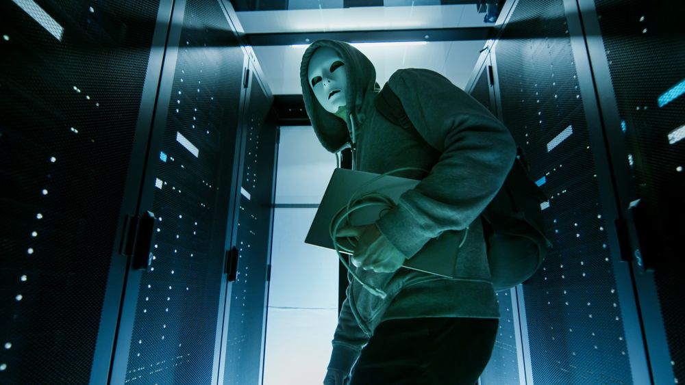 Low Angle Shot of a Masked Hacker in a Hoodie Sneaking Through Corporate Data Center with Rows of Rack Servers.