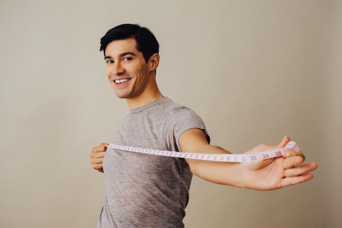 Smiling Hispanic male holding measuring tape out in front of him in beige studio shoot