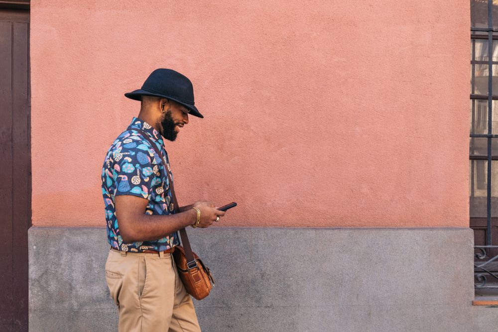 Stylish Black man walking on the city street using smartphone