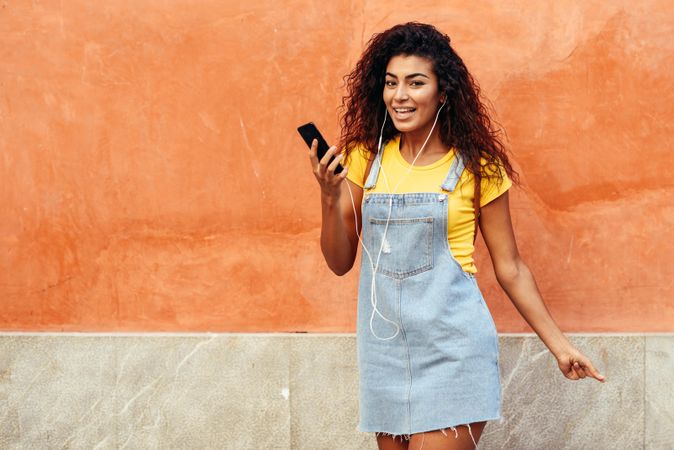 Happy Arab woman with curly hairstyle using phone and headphones in front of on orange wall