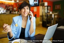 Happy woman sitting in cafe speaking on phone with laptop and slice of cake 0LdERR