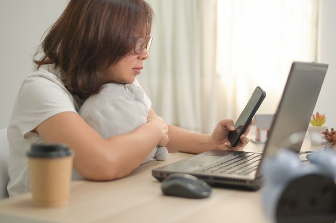 A woman holding a smartphone and using a laptop working from home, home office.