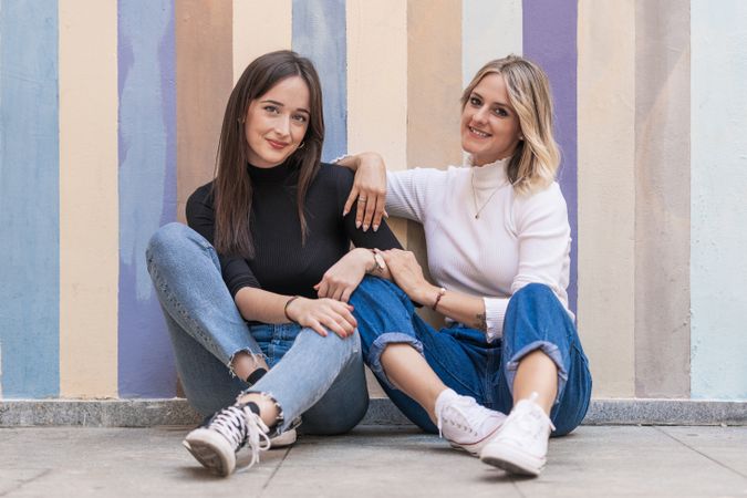Happy girlfriends sitting against painted urban wall