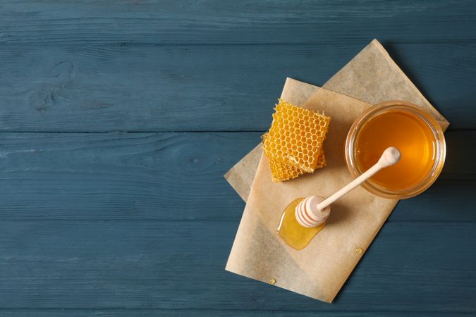 Glass jar with honey, dipper, honeycombs and parchment on wooden background, copy space