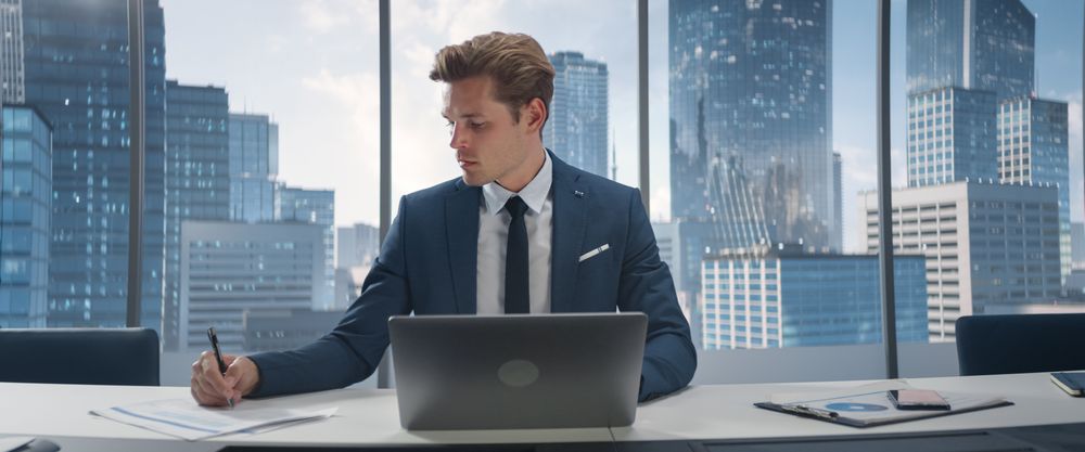 Confident Businessman in a Suit Sitting at a Desk in Office, Using Laptop Computer, Next to Window with Big City with Skyscrapers View.