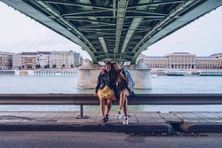 Smiling girlfriends hugging and sitting on fence along river under bridge