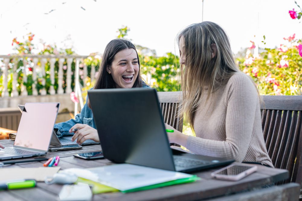 Two smiling female friends comparing their notes and chatting about what was written on the laptop.